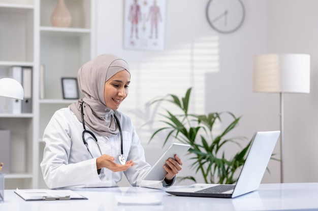 Arab muslim woman doctor sits at a table in the office in front of a laptop conducts an online