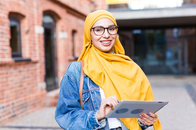 Arab woman student. Beautiful muslim female student wearing bright yellow hijab holding tablet.