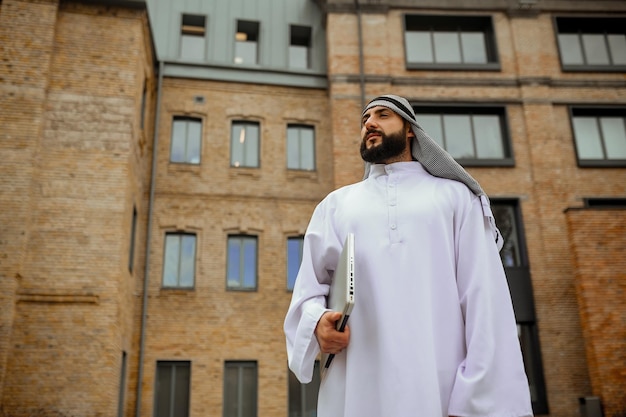 An arabian man in traditional clothing with a laptop in the street