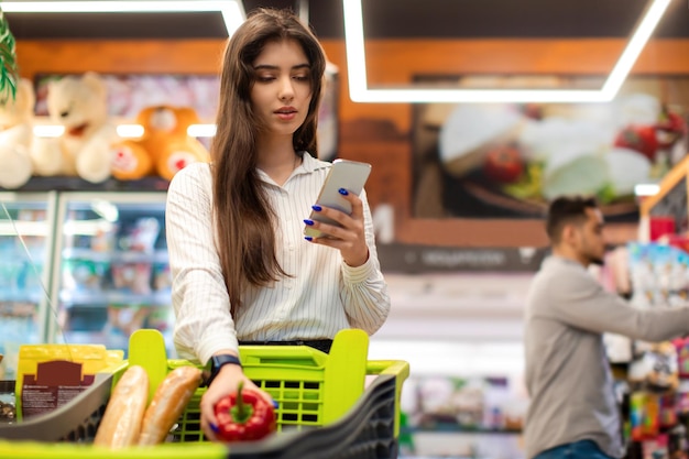 Arabic young woman using smartphone application shopping groceries in supermarket