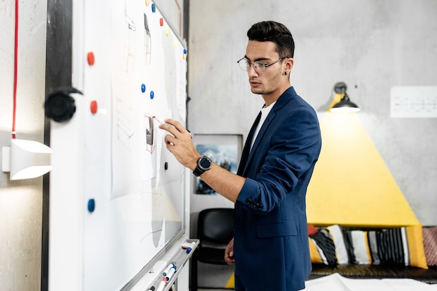 The architect dressed in blue checkered jacket attaches blueprints to a magnetic board on the wall in the office .