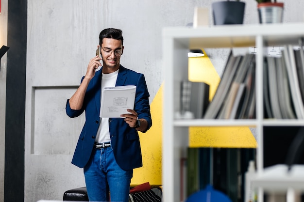 Architect dressed in stylish clothes talking by phone in a room with a stylish interior with bookshelves .