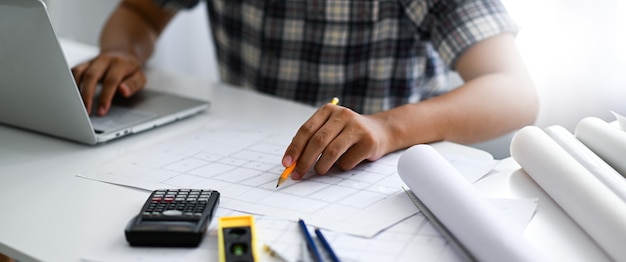 The architect is pointing a pencil at a house plan and working on a laptop. He is checking and recording the results, calculators, and drawings on the desk.