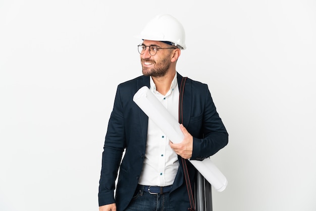 Architect man with helmet and holding blueprints isolated on white background looking to the side and smiling