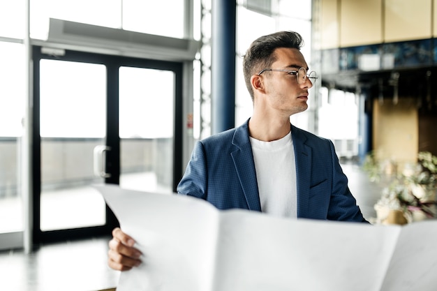 Architect in stylish clothes holds sheet with drawing in his hand and talks by phone on the background of a modern glass multistory building .