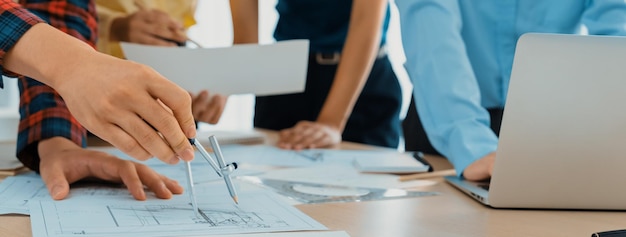 Architect team discuss about blueprint design at modern meeting room while her coworker working on laptop on table with blueprint and architectural equipment scattered around Closeup Delineation