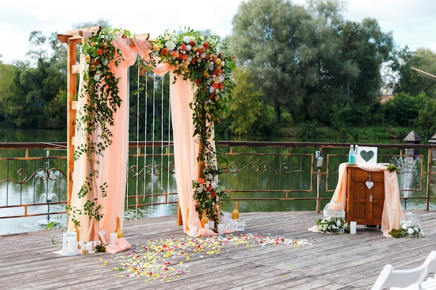 Area of the wedding ceremony near river on the pier. Wooden rectangular arch, white chairs decorated with flowers, greenery, petals, leaves and tulle in peach color. Cute, trendy rustic decor