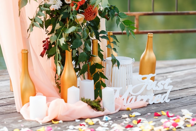 Area of the wedding ceremony near river on the pier. Wooden rectangular arch, white chairs decorated with flowers, greenery, petals, leaves and tulle in peach color. Cute, trendy rustic decor