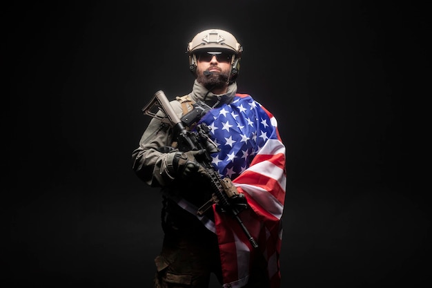 Photo army of america a soldier in military equipment with a gun holds the usa flag on a black background