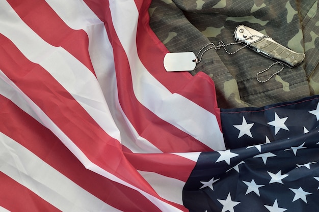 Army dog tag token and knife laying on camouflage uniform and folded United States flag