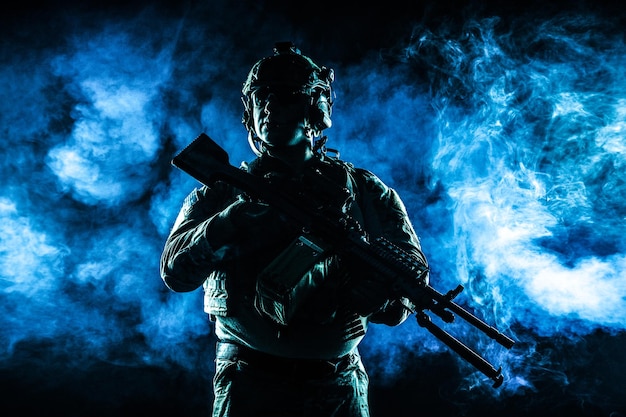 Army soldier in Combat Uniforms with machine gun, plate carrier and combat helmet are on. Studio contour silhouette shot, backlight, dark glowing smoke background