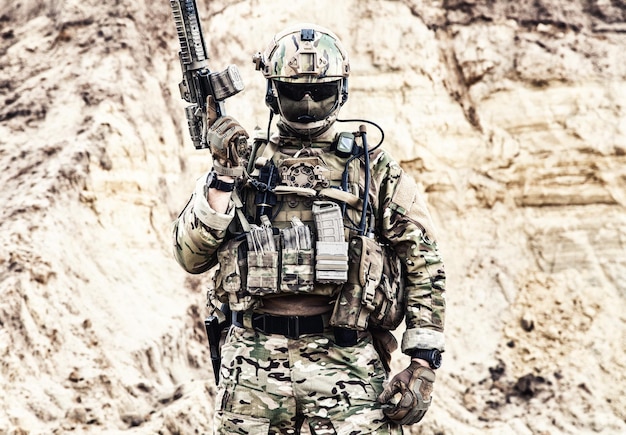 Army soldier, private military company fighter with hidden identity in battle uniform, helmet, body armor and radio headset, standing with service rifle in rocky area