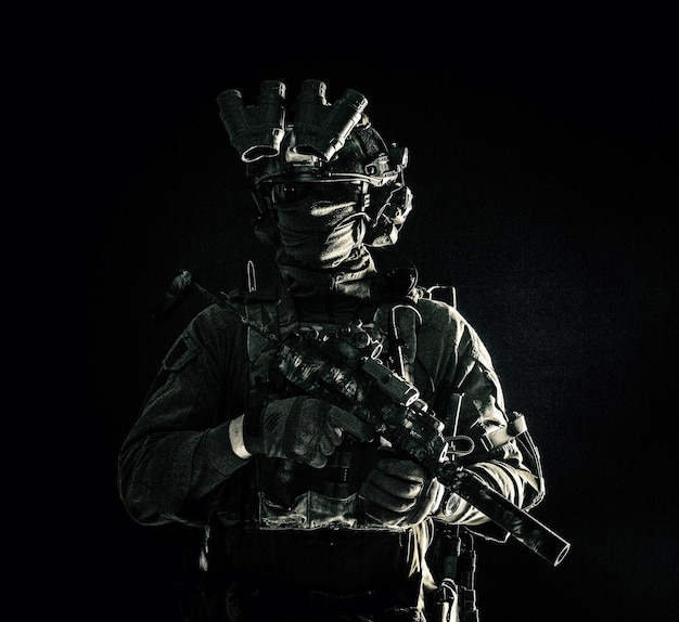 Army special operations forces soldier in mask and combat uniform helmet equipped nightvision device armed submachine gun with silencer looking aside low key studio portrait on black copyspace