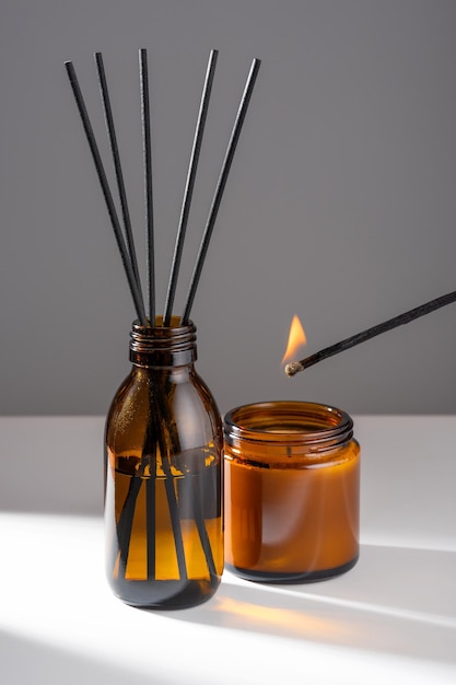 Aroma diffuser with natural essential oil in brown glass bottle soy and coconut wax candle with fire lit by a match in a Amber jar on a white gray background Concept mental health and harmony