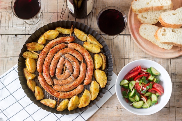 Aromatic meat sausages with potatoes, salad and wine on a wooden table.