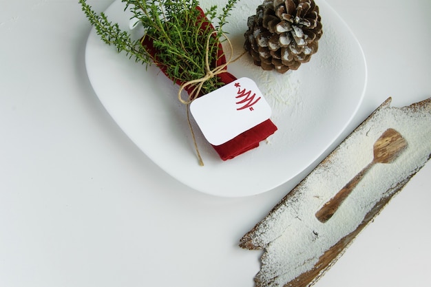 Arranging the table for the winter holidays concept. Table decoration in minimal style, red napkin with herbs, the outline of a dessert spoon, pine cone and holiday place card