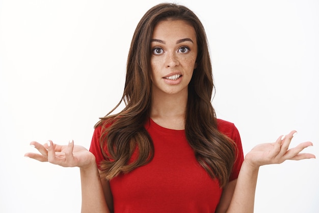 Arrogant snobbish young brunette in red t-shirt shrugging raise hands look with disdain and confusion, feeling questioned and bothered with strange accusation, white wall