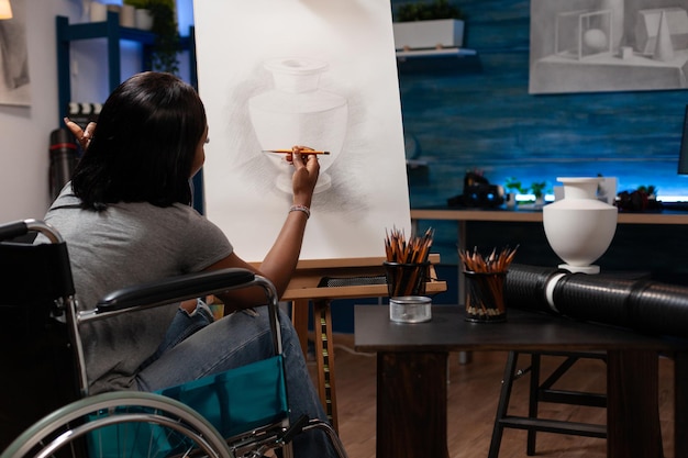 Art student in wheelchair sketching vase on canvas working at artistic ilustration using grahic pencil during painting lesson in creativity home studio. Young woman disability with drawing sketch