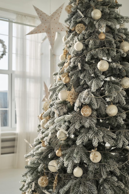 Artificial lush cast snowy Christmas tree spruce decorated with white golden balls candles lights garland close up in white interior