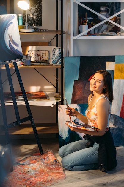 Artist lifestyle Painting craft Creative home leisure Happy satisfied smiling painter woman sitting on floor with abstract picture on easel in cozy home studio