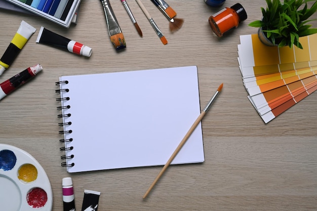 Artist workspace with empty notebook surrounded by color swatches, paintbrushes and other various equipment on wooden background.