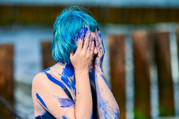 Artistic blue-haired woman performance artist in dark blue dress smeared with indigo gouache painting on her body on beach, close up. Creative body painting, body art concept, outdoor performance art