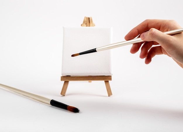 Artists hand with paintbrush painting making stroke on blank empty square canvas on mini easel