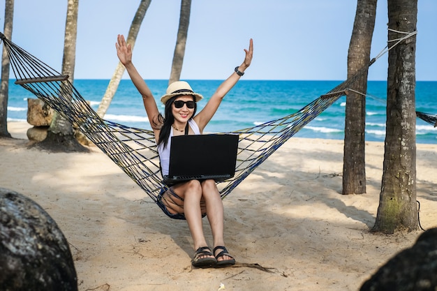 Asia woman sitting on the hammock near the beach and using laptop for her work and checking business during her vacation.