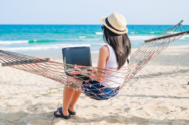 Asia woman sitting on the hammock near the beach and using laptop for her work and checking business during her vacation.