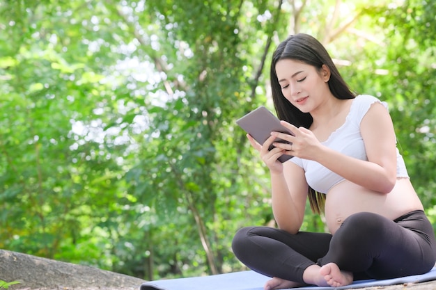 Asian 8 month pregnant woman using digital tablet sitting on grass in outdoor park. Outdoor nature background.