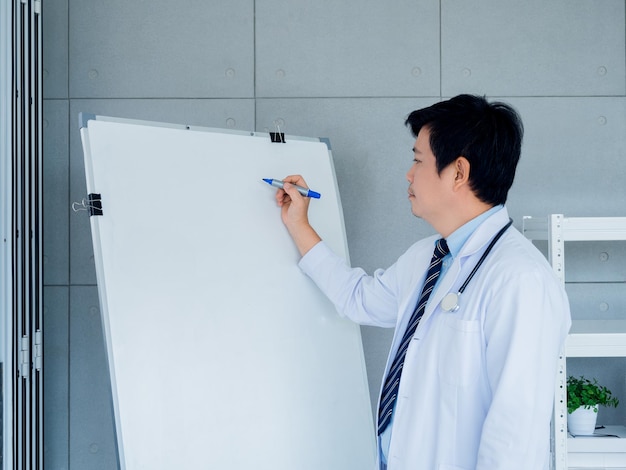 Asian adult male doctor in white uniform with stethoscope is holding chemical pen writing on a blank whiteboard to describe a patient or practitioner students in medical office in hospital room