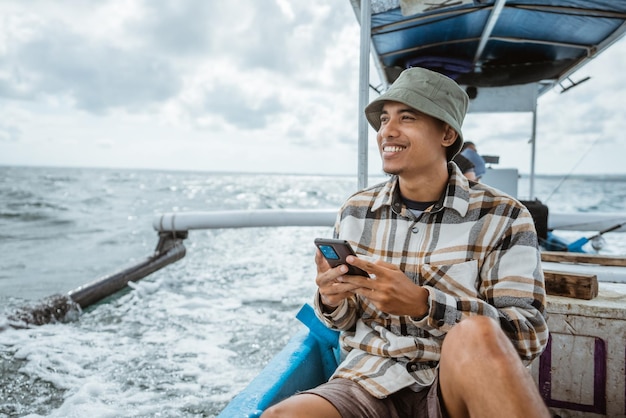 Asian angler sits relaxed while using smartphone on fishing boat