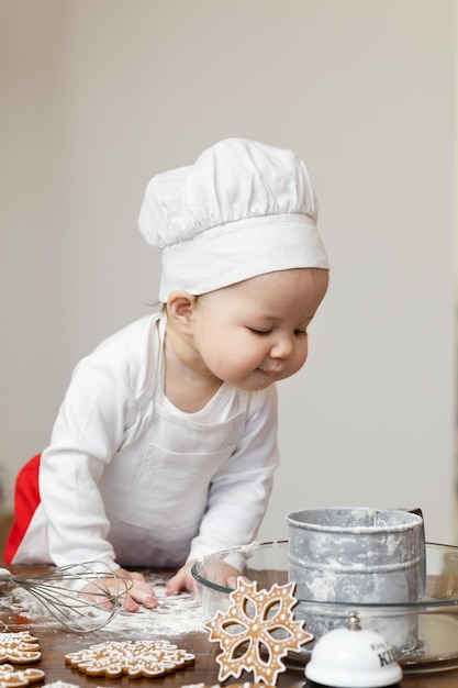 An Asian baby in a chef's hat and apron is preparing Christmas ginger cookies With curiosity he looks into the container with flour