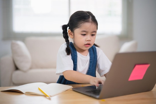 Asian baby girl smiling use laptop or writing drawing color on note book study online on wood table desk in living room at home Education learning online from home concept