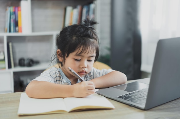 Asian baby girl wearing a blue striped shirt use laptop and write notes in notebook to study online on wood table desk in living room at home Education learning online from home concept