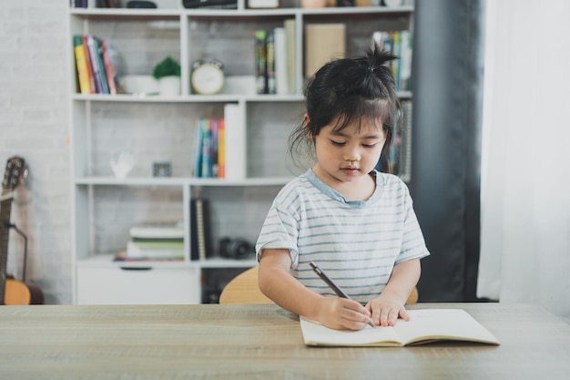 Asian baby girl wearing a blue striped shirt write notes in notebook to study online on wood table desk in living room at home Education learning online from home concept