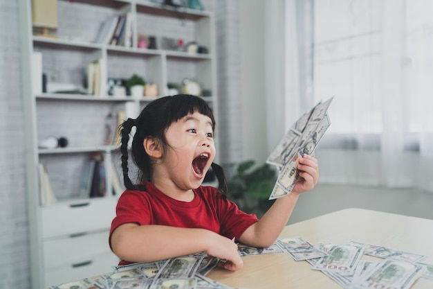 Asian baby girl wearing a red tshirt holding dollar bill on wood table desk in livingroom at home Saving investment wealth concept