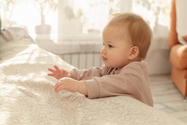 An Asian baby stands in the bedroom leaning on the bed and trying to take her first steps