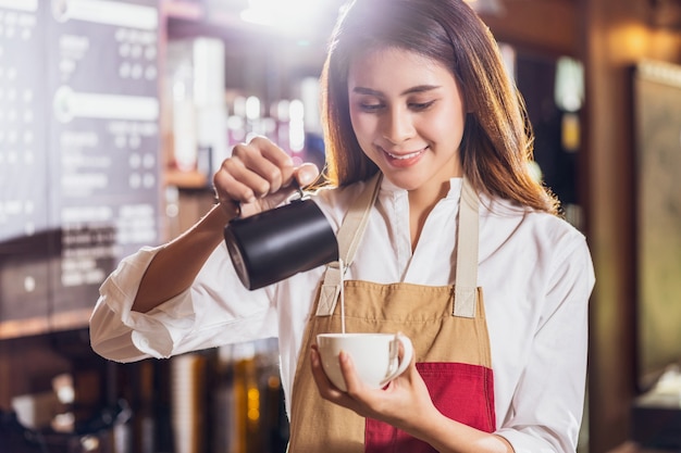 Asian Barista pouring milk to cup of coffee which is espresso with latte or cappuccino for customer