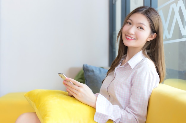 Asian beautiful girl with long hair who wears shirt sits on yellow sofa while she plays smartphone and smile happily in working space.