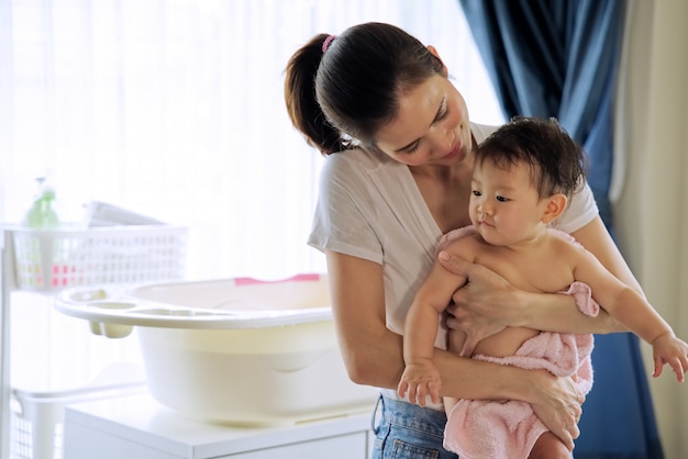 Asian beautiful mother holding little cute baby after taking a bath and looking and her child standing in the room