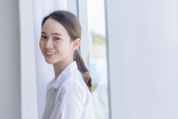 Asian beautiful woman has black long hair in white shirt She is smiling and standing near window