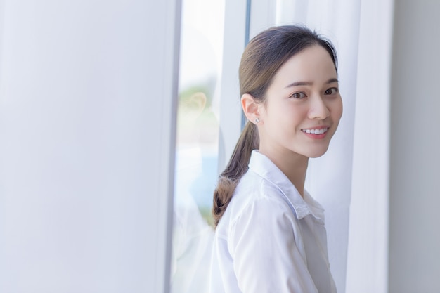 Asian beautiful woman in white shirt is standing lean against with window and white curtain.