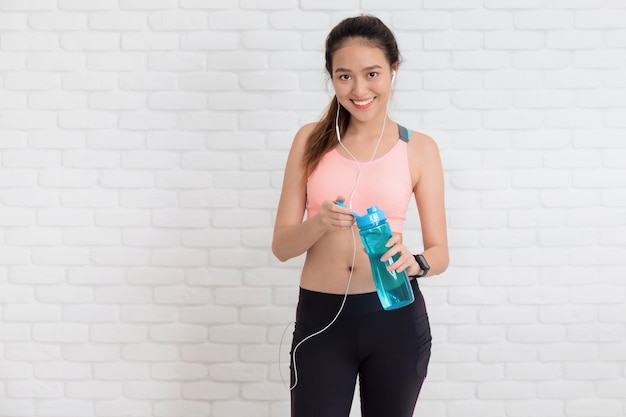 Asian beautiful women holding water bottle after play yoga and exercise on white brick wall background with copy space.Exercise for Lose weight, increase flexibility and tighten the shape.