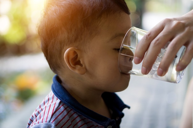 Asian boy is drinking water from the clear glass entered by his mother.