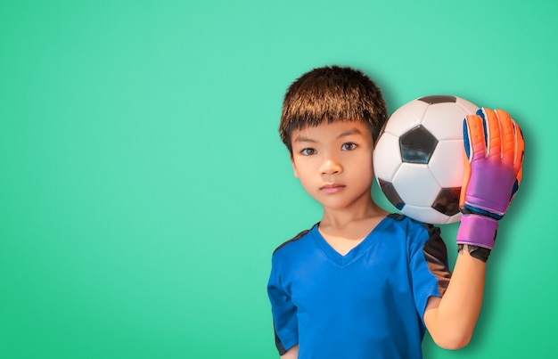 Asian boy is a football goalkeeper wearing gloves and holding a soccer ball on green background copy space.