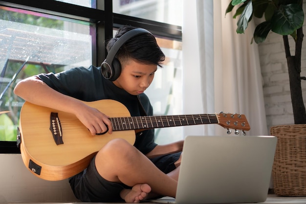 Asian boy playing guitar and watching online course on laptop while practicing for learning music