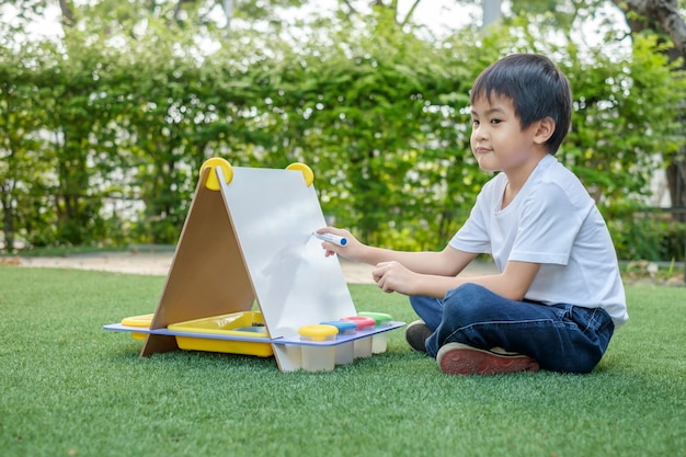 An Asian boy in a white t-shirt and jeans is sitting outside drawing on a whiteboard in the grass.