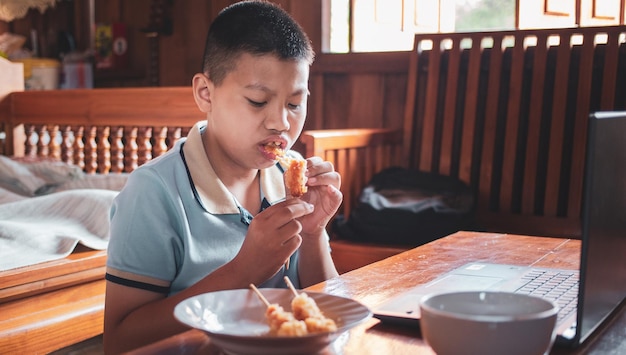 Asian boys eat junk food while sitting at a desk with notebooks and laptops at home