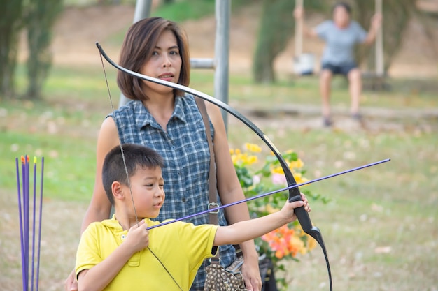 Asian boys holding a bow in camp adventure and the woman standing behind Background  blurry tree.